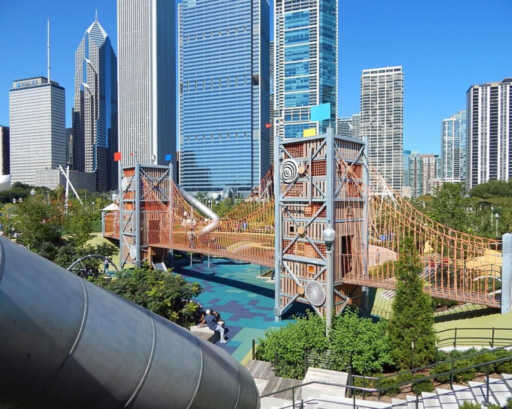 Suspension bridge structure in an urban park with tall office buildings in the background, blue sky above.