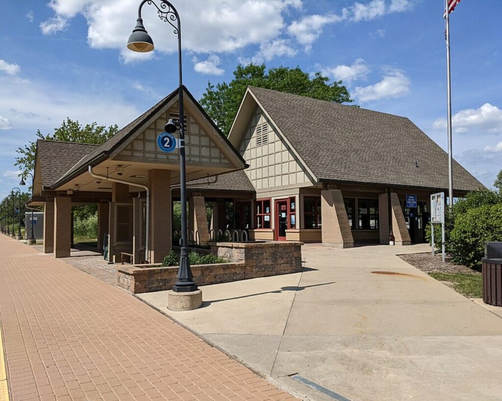 Small commuter train station with a covered platform, brick walkway, and a blue '2' sign under a decorative street lamp on a sunny day