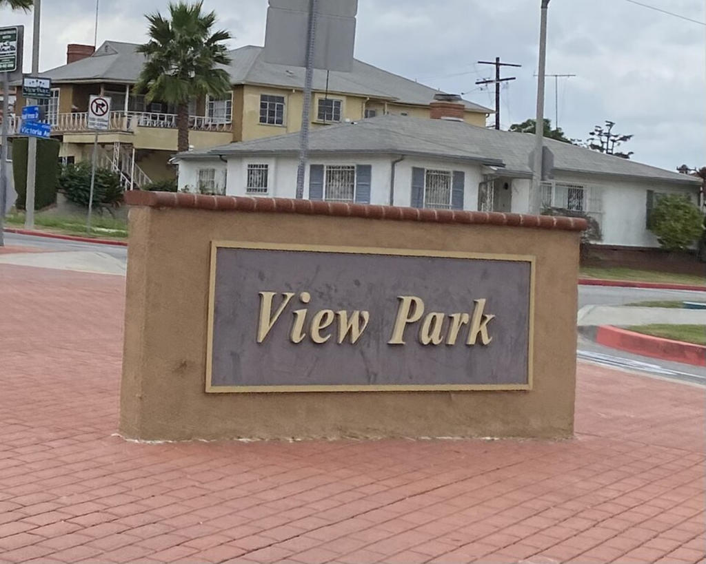View Park neighborhood sign in Los Angeles, set on a brick-paved corner with nearby homes, palm trees, and street signs.