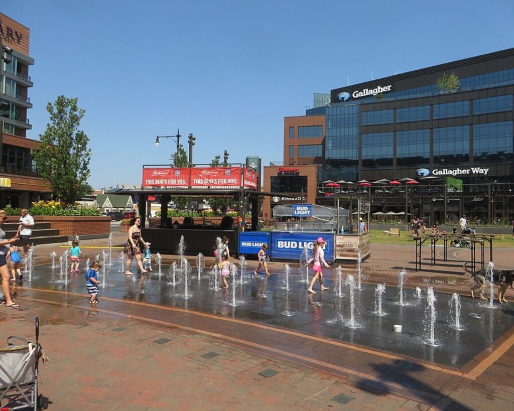 Children and adults play in a sunny splash fountain at a outdoor plaza with Bud Light kiosks and a glass-fronted building in the background, labeled Gallagher Way/Gallagher Way.