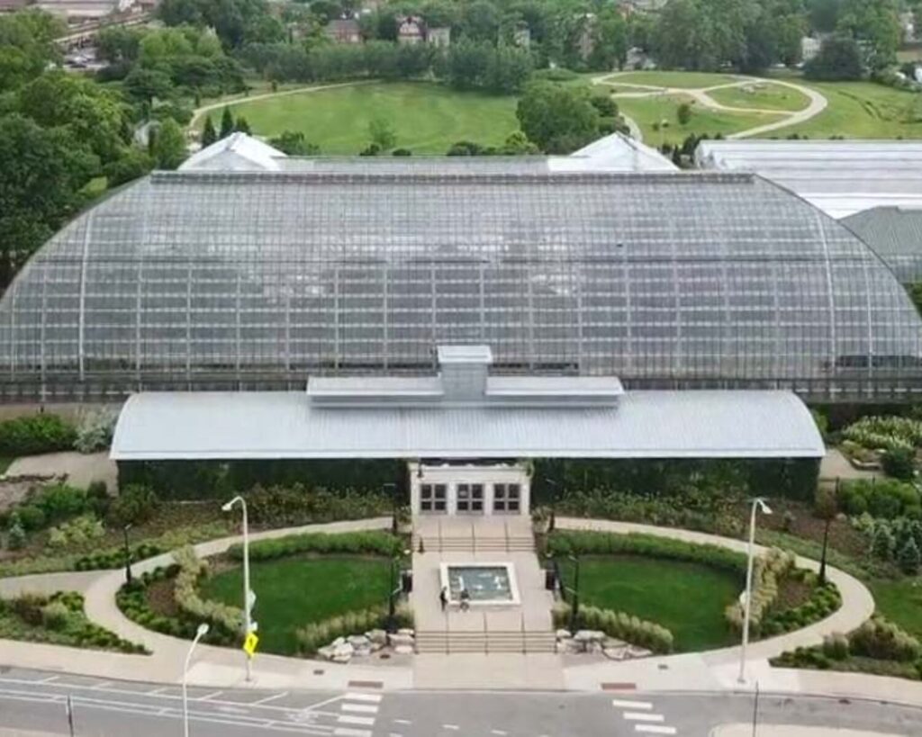 Large glass greenhouse with curved roof and central entrance, surrounded by manicured gardens and a sidewalk.