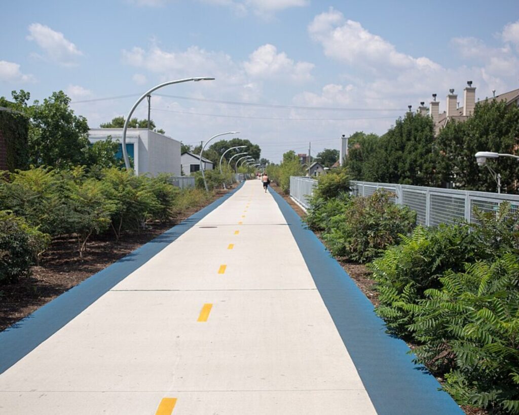 Long white bike/pedestrian path with blue borders, curved light poles, and greenery on both sides; a cyclist in the distance.