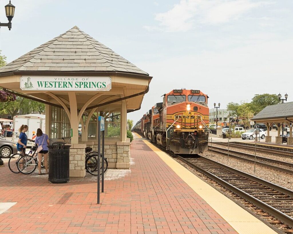 Orange diesel locomotive arrives at the Western Springs station as passengers with bicycles stand near the platform shelter.