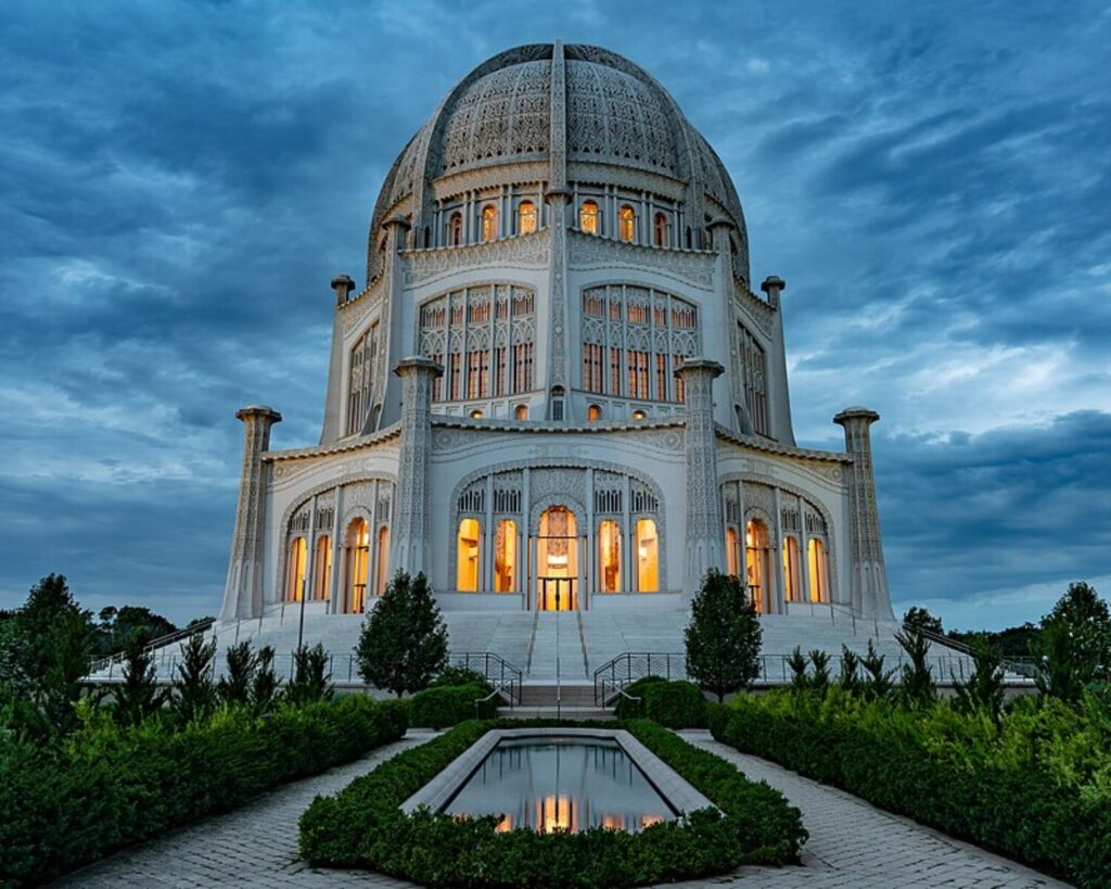 White ornate domed temple at dusk with glowing windows and intricate facade, surrounded by green hedges and a reflecting pool in the foreground.