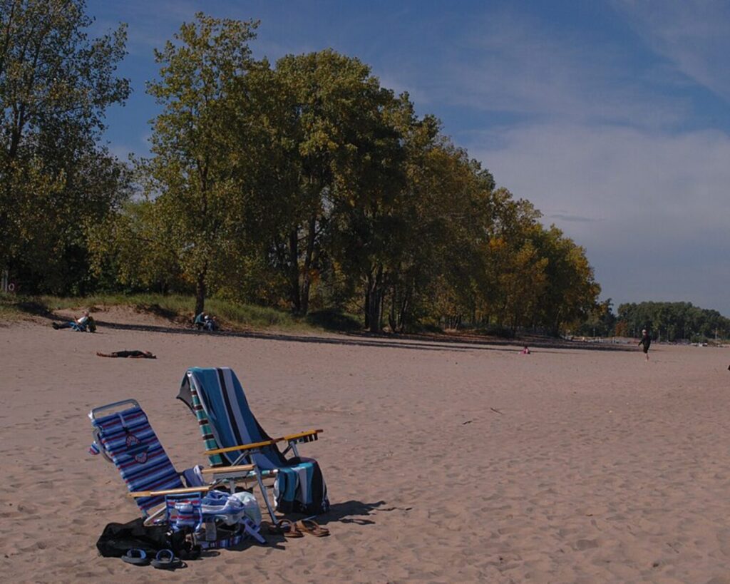 Two blue striped beach chairs sit on the sandy shore with a line of trees in the background and a few people relaxing nearby.