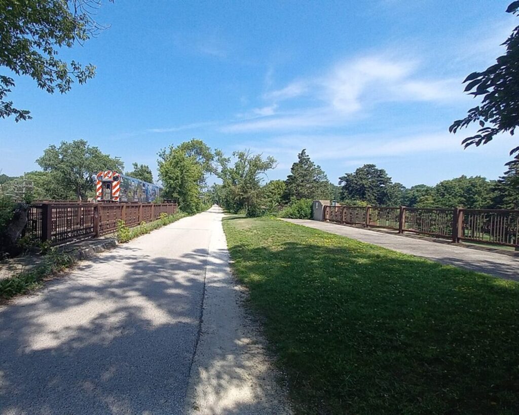 Green Bay Trail crossing over Winnetka Avenue beside the Metra Union Pacific North railway, with a train passing through a tree-lined setting.