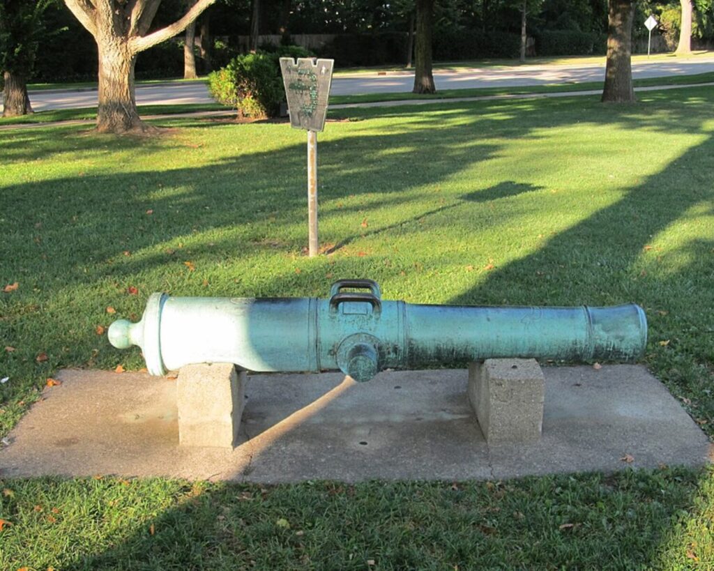 Decorative green cannon mounted on concrete blocks in a sunny park lawn with a sign nearby