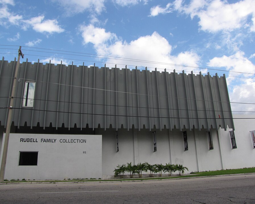 Rubell Family Collection building in Miami, featuring a modern gray-and-white facade with narrow windows and palm landscaping beneath a partly cloudy sky.