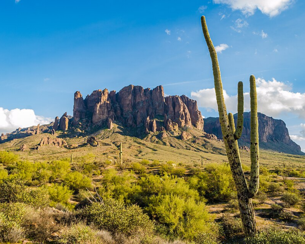 Superstition Mountains near Apache Junction, Arizona, with towering rocky cliffs, saguaro cacti, desert shrubs, and a bright blue sky.