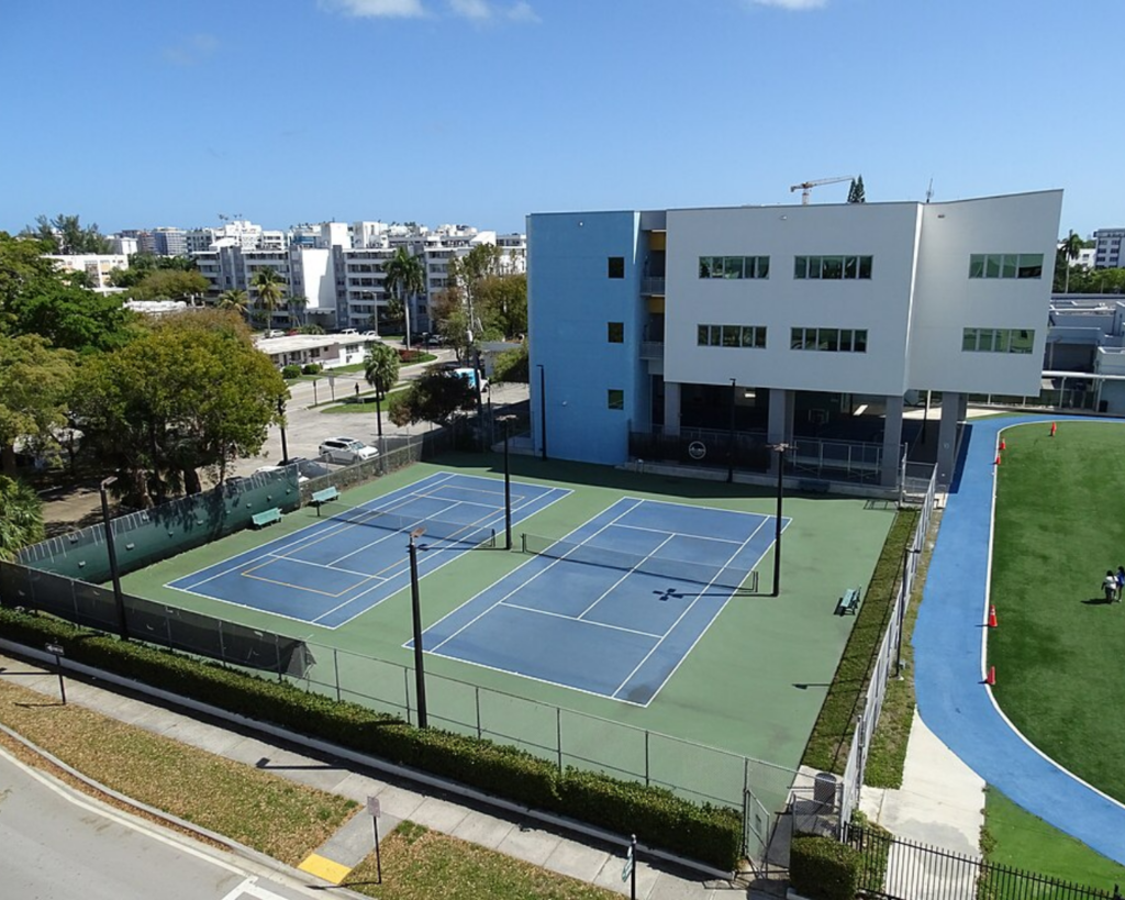 Tennis courts in Bay Harbor Islands, Florida, beside a modern building, athletic field, and surrounding residential buildings under a clear blue sky.