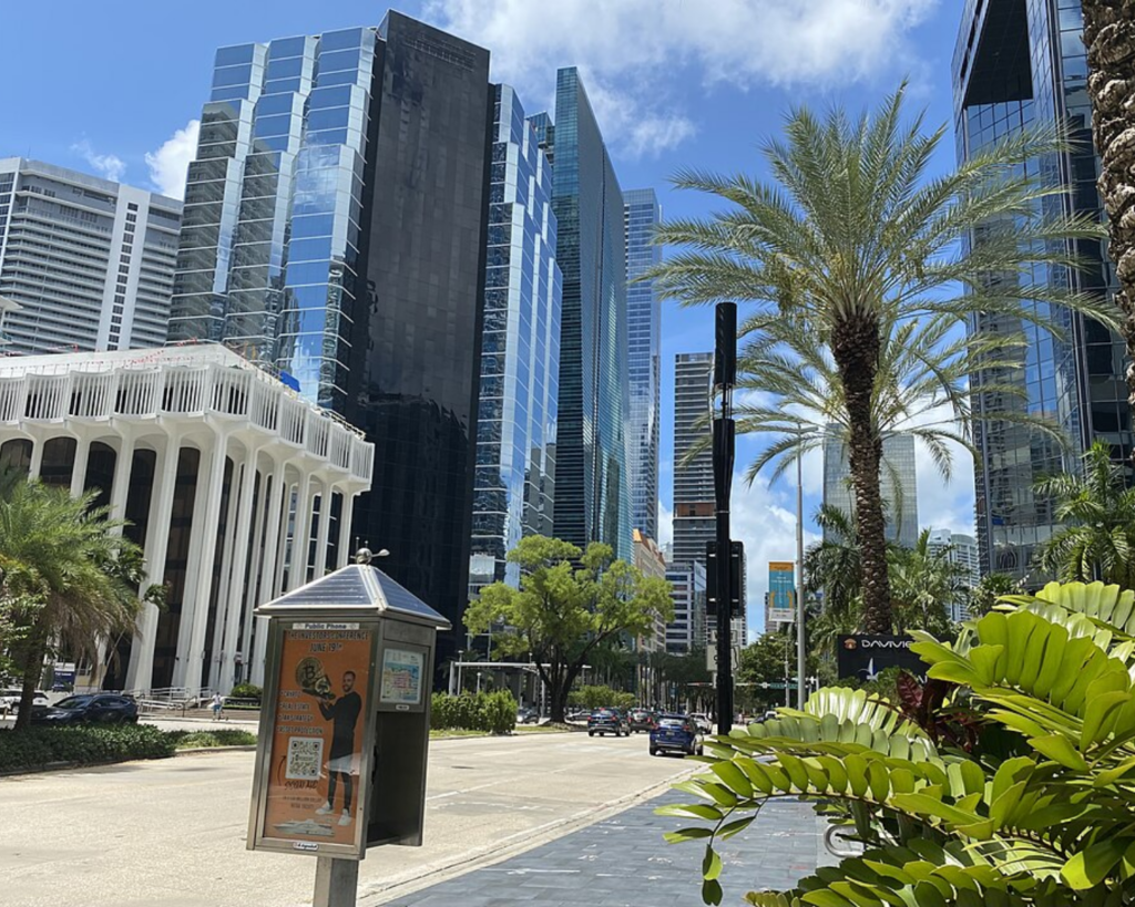 Street view of Brickell in Miami, Florida, with modern glass skyscrapers, palm trees, traffic, and a public pay phone kiosk in the foreground.