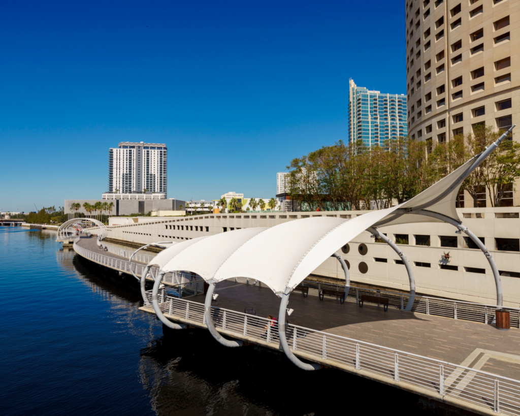 Tampa Riverwalk in Florida, featuring a waterfront promenade with white canopy structures, calm blue water, and high-rise buildings under a clear blue sky.