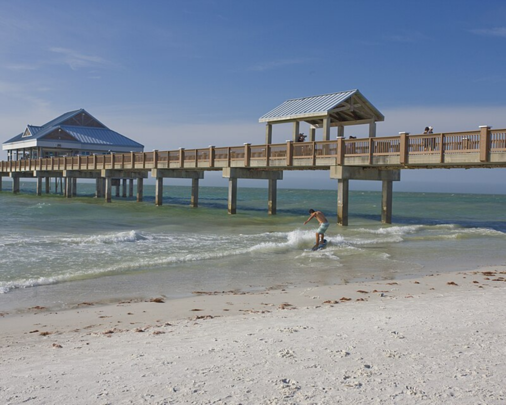 Clearwater Beach, Florida, with a long fishing pier extending over turquoise Gulf waters, gentle waves, and a white sandy shoreline under a clear blue sky.