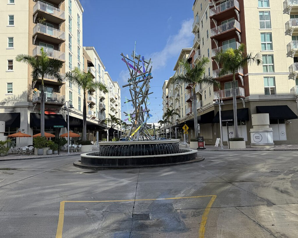 Downtown Dadeland in Miami, Florida, with palm-lined apartment buildings, outdoor dining, a circular fountain, and a modern abstract sculpture.