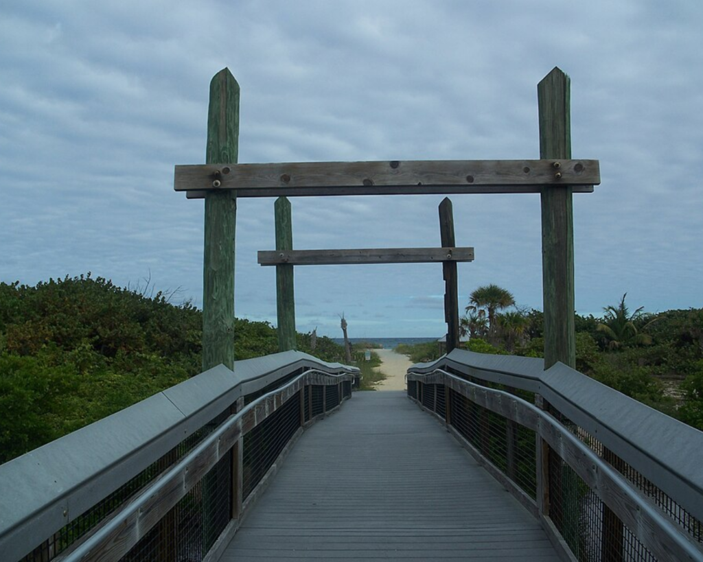 Wooden boardwalk at John U. Lloyd Beach State Park in Dania Beach, Florida, leading through coastal vegetation toward the ocean under a cloudy sky.