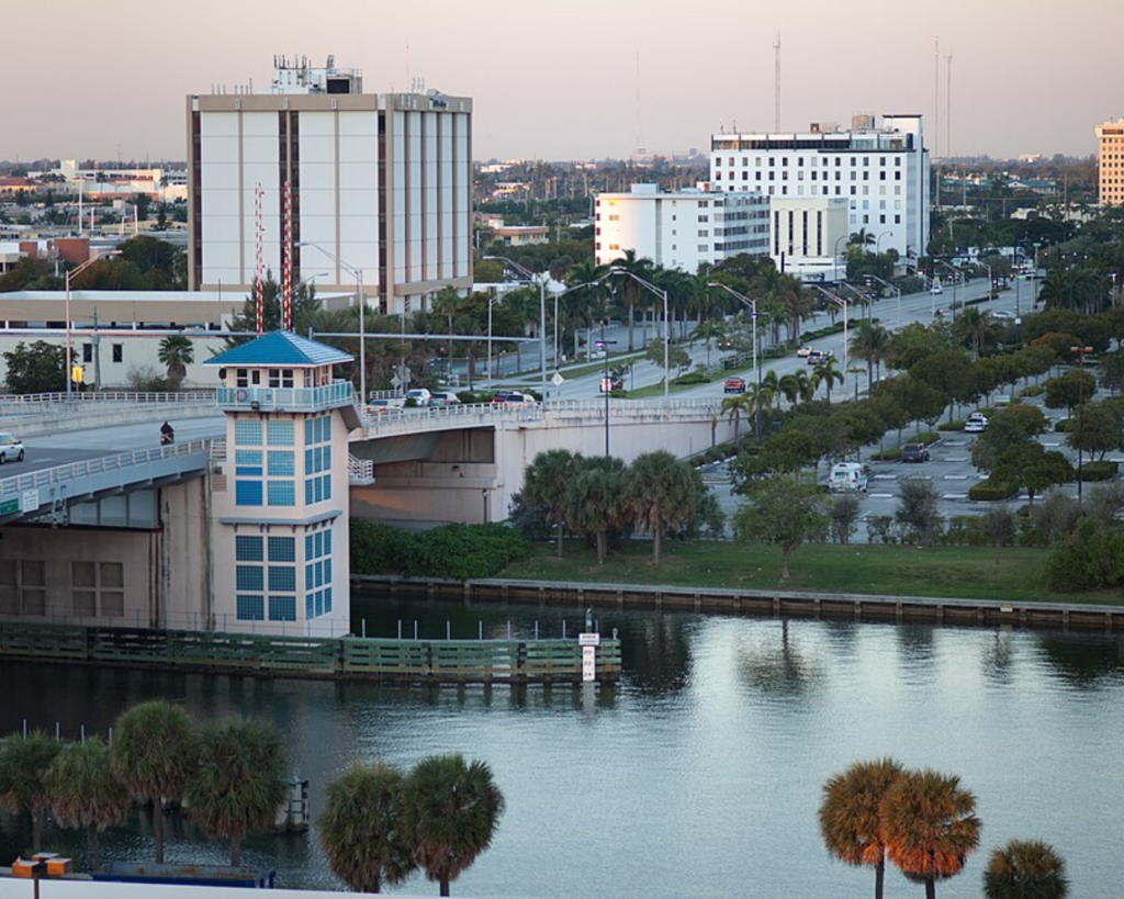 Hallandale Beach Boulevard Bridge over a waterway in Hallandale Beach, Florida, with palm trees, roads, and mid-rise buildings in the background.