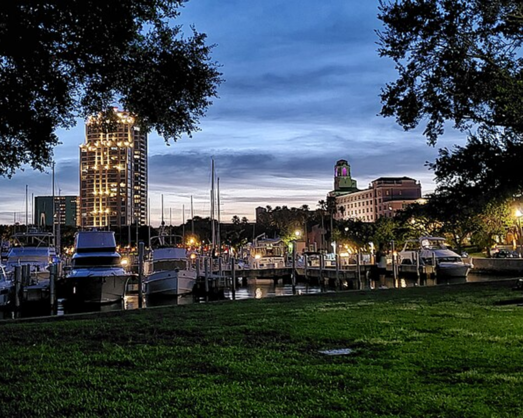 Vinoy Park in downtown St. Petersburg, Florida, at dusk, with a marina full of boats, illuminated high-rise buildings, green lawn, and trees framing the waterfront.