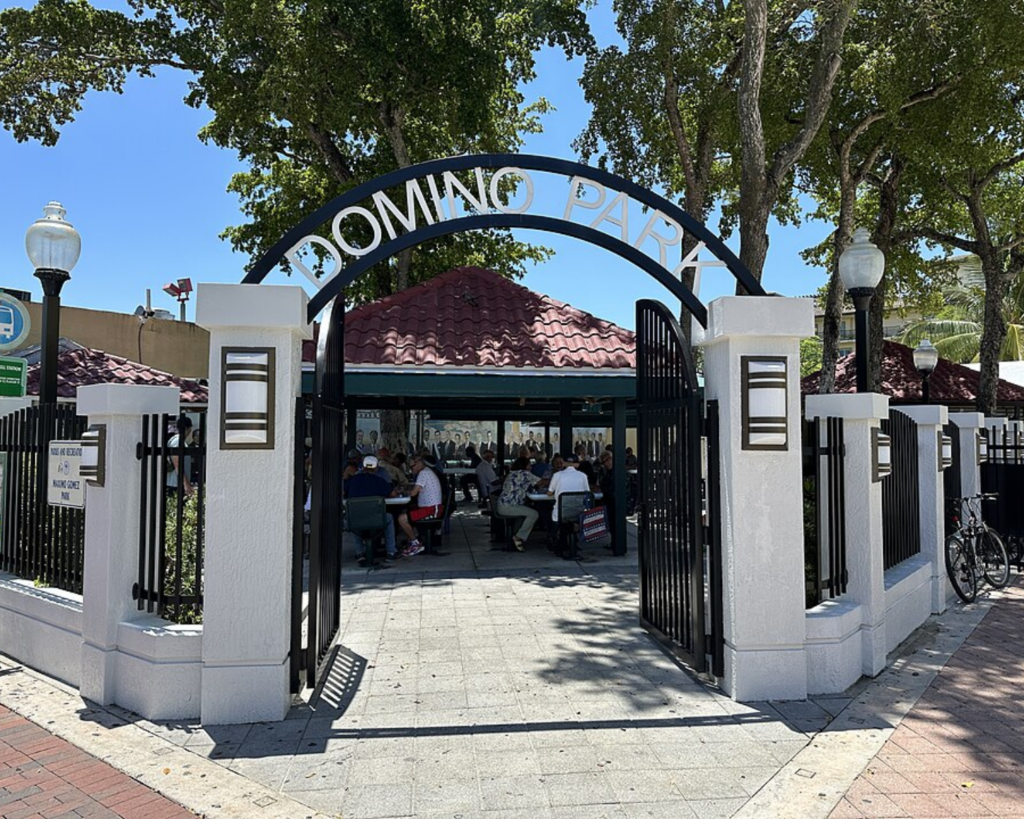 Entrance to Domino Park in Miami’s Little Havana, with visitors seated at tables beneath shaded pavilions and large trees.