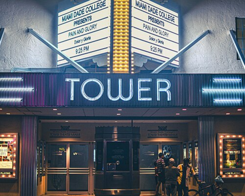 Night view of the historic Tower Theater in Miami’s Little Havana, with its illuminated Art Deco marquee, entrance, and people gathered outside.