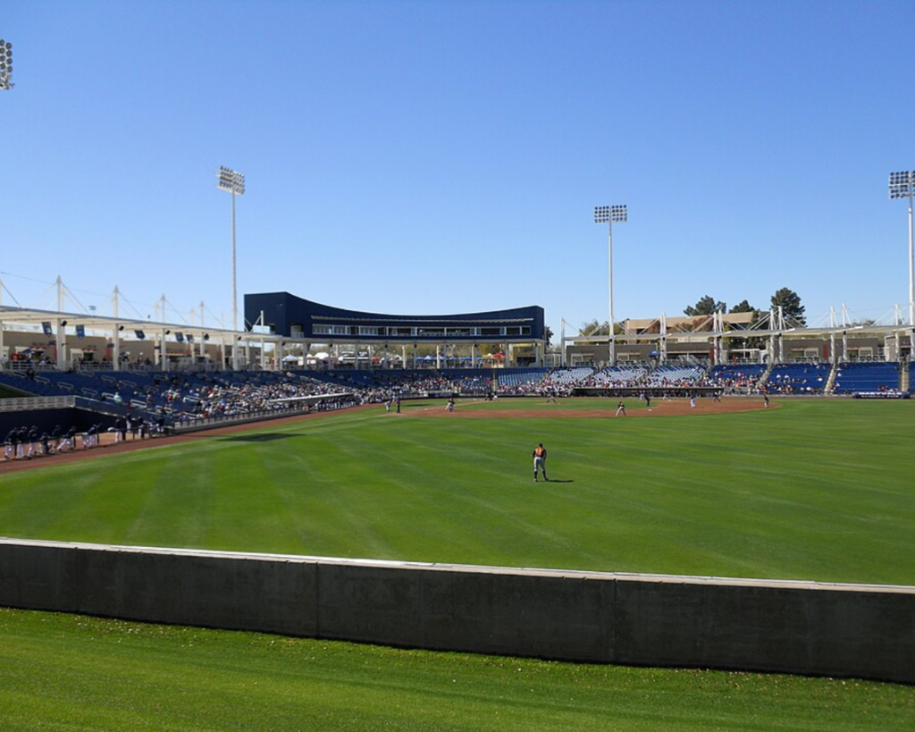 Maryvale Baseball Park in Phoenix, Arizona, showing the green baseball field, stadium seating, spectators, and tall floodlights under a clear blue sky.