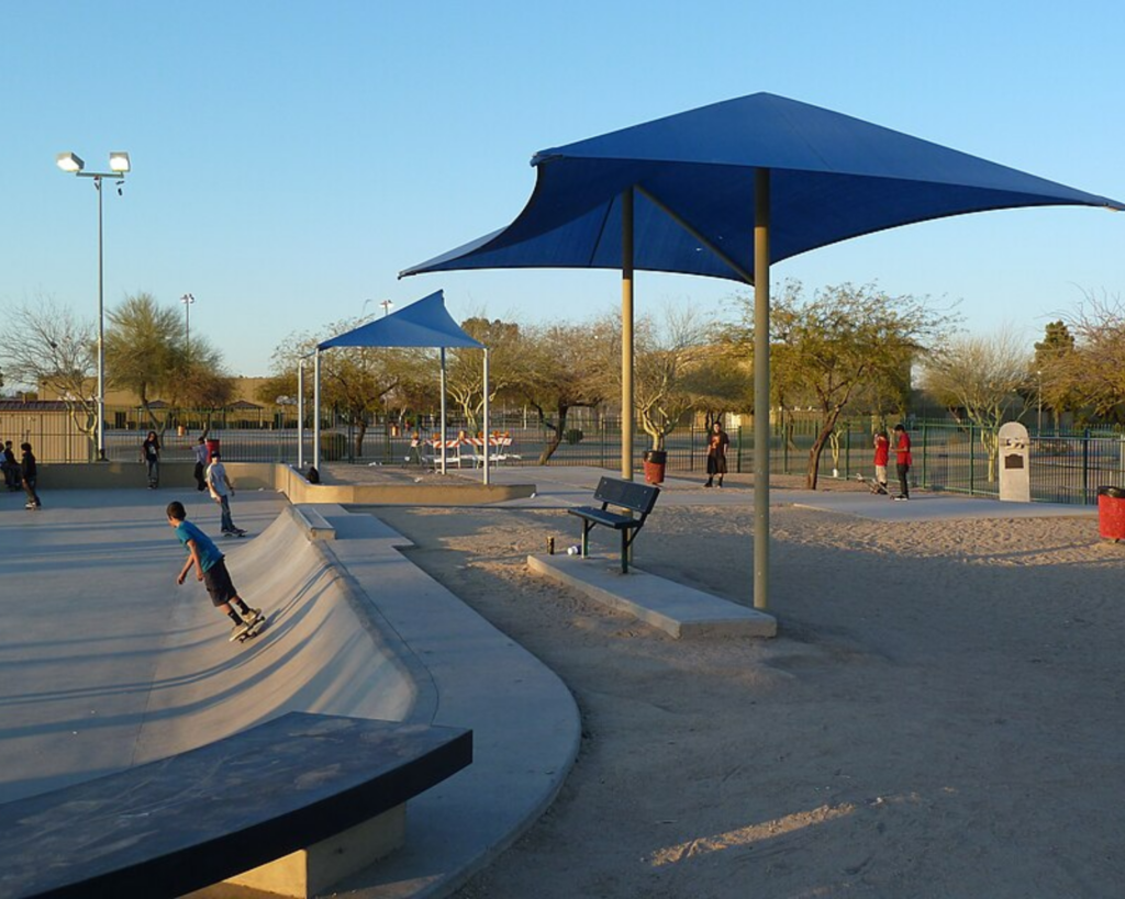Desert West Community Center Skateboard Plaza in Maryvale, Phoenix, Arizona, with skateboarders, concrete ramps, benches, and large blue shade canopies.