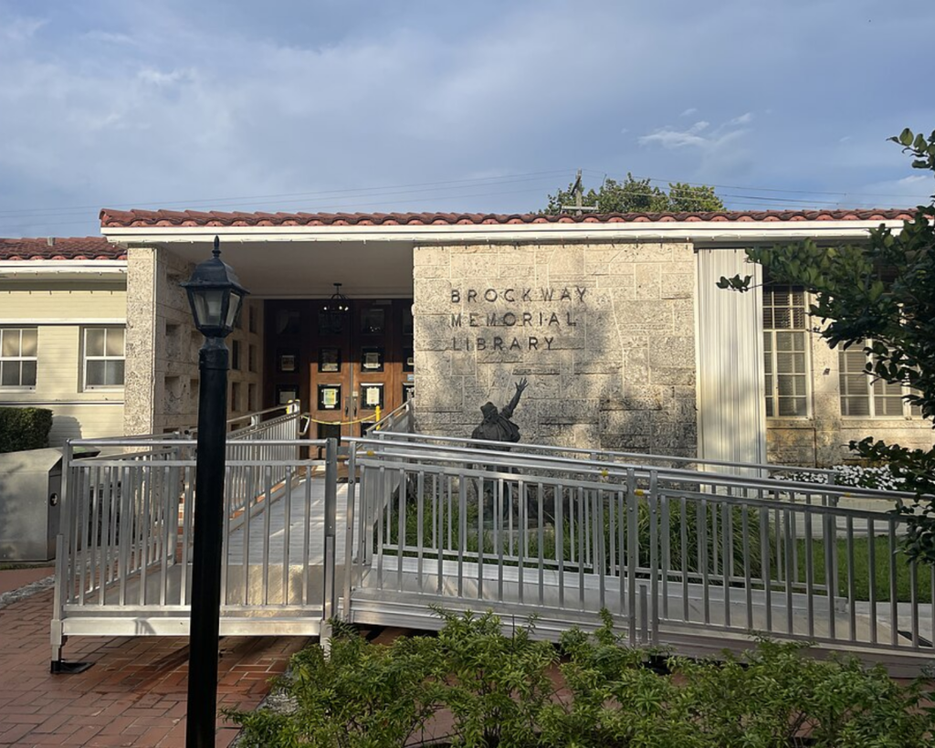 Brockway Memorial Library in Miami Shores, Florida, showing its stone-front entrance, accessibility ramp, landscaped grounds, and a black lamppost in front.