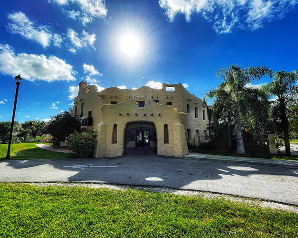 Curtiss Mansion in Miami Springs, Florida, featuring a distinctive Pueblo Revival-style facade, palm trees, green lawns, and a bright blue sky.