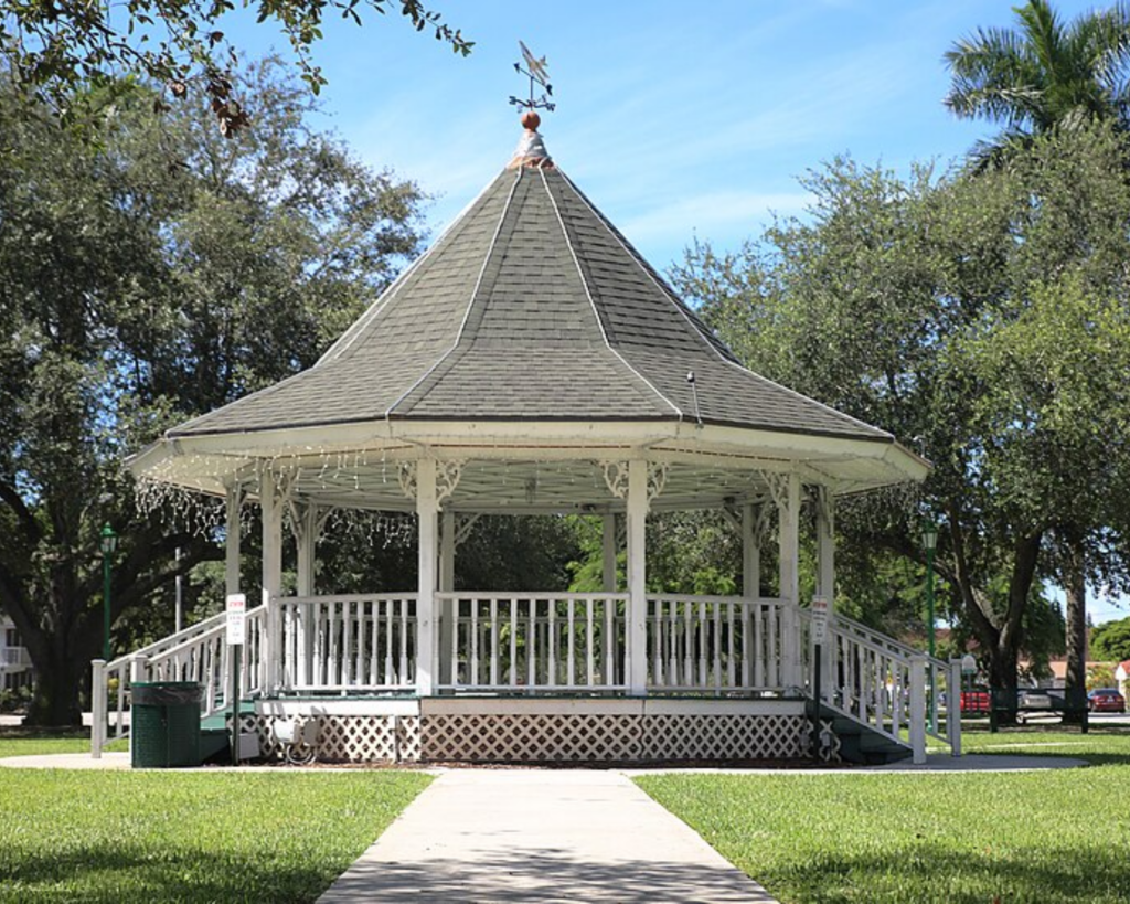 White gazebo at Glenn H. Curtiss Memorial Circle in Miami Springs, Florida, surrounded by green lawn, trees, and palm trees under a clear blue sky.