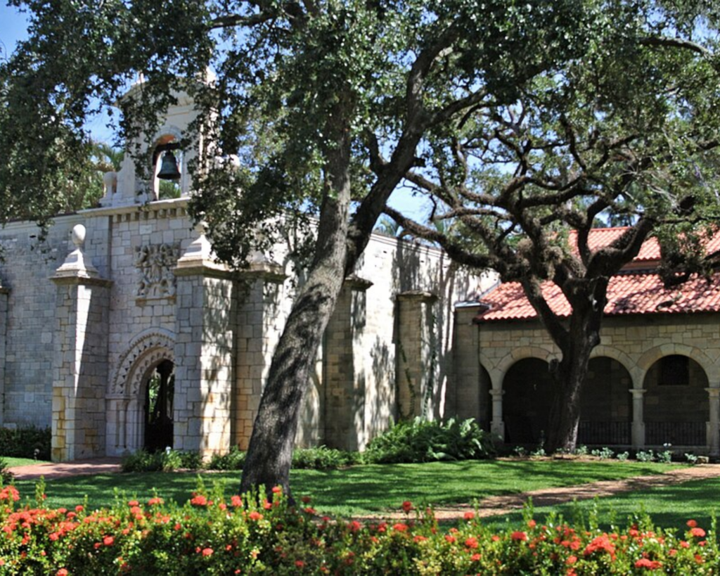 St. Bernard de Clairvaux Church in North Miami Beach, Florida, with a stone facade, bell tower, red-tiled roof, large shade trees, and colorful flower beds.