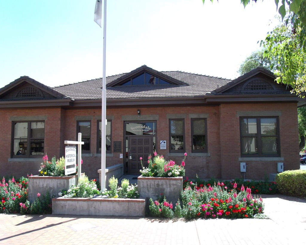 Historic Scottsdale Grammar School building in Scottsdale, Arizona, featuring a red-brick facade, dark wood trim, landscaped flower beds, and a flagpole in front.