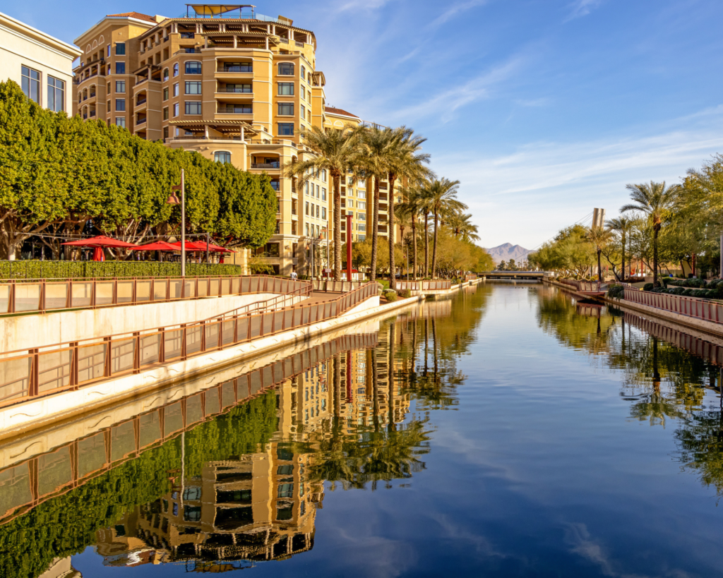 Scottsdale Waterfront in Arizona, showing a calm canal lined with palm trees, landscaped walkways, and multi-story buildings reflected in the water.