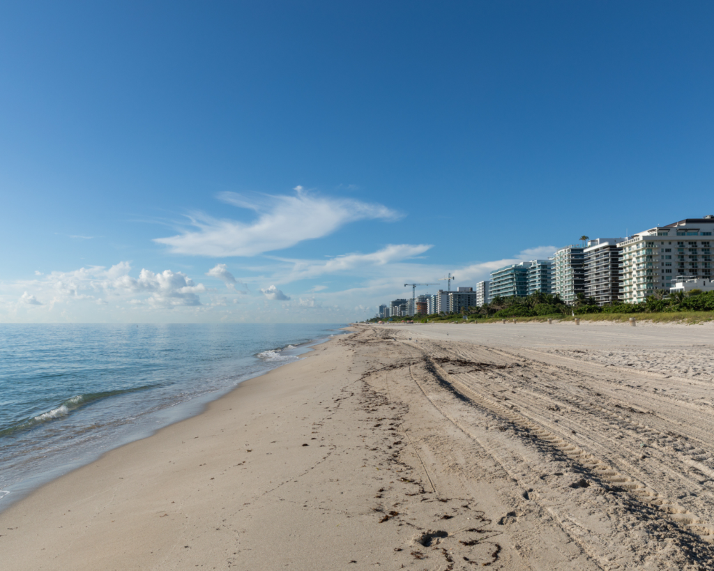 Surfside Beach in Florida, showing a wide sandy shoreline, calm Atlantic Ocean, and beachfront high-rise buildings beneath a clear blue sky.