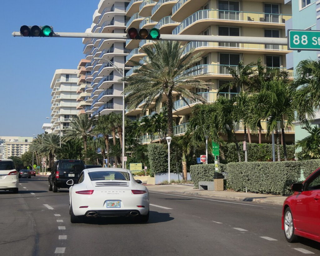Street view in Surfside, Florida, with palm trees, high-rise apartment buildings, cars, and traffic lights along a sunny boulevard.