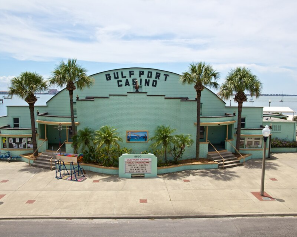 Gulfport Casino in Gulfport, Florida, featuring a pale green waterfront building, palm trees, paved plaza, and calm bay waters in the background.