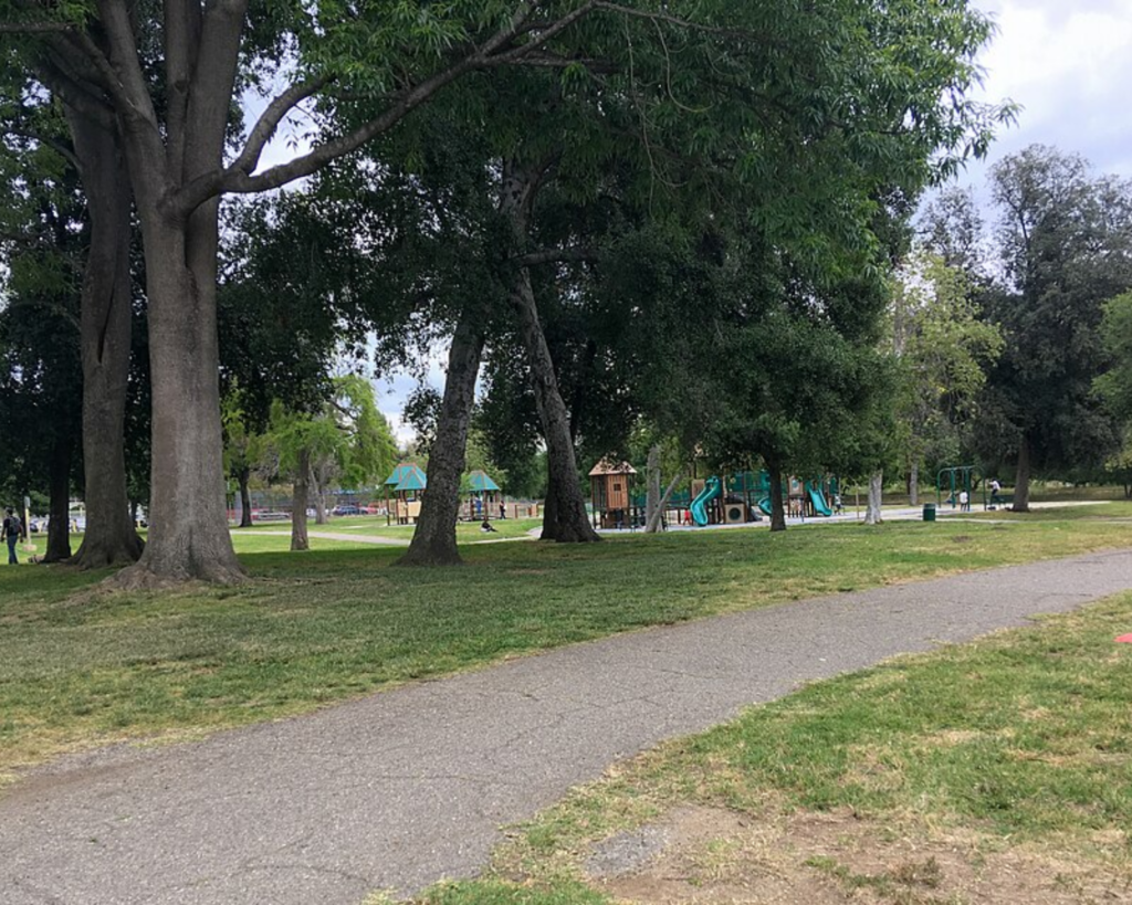 Sycamore Grove Park in Los Angeles, California, featuring large shade trees, grassy lawns, a paved walking path, and a playground in the background.