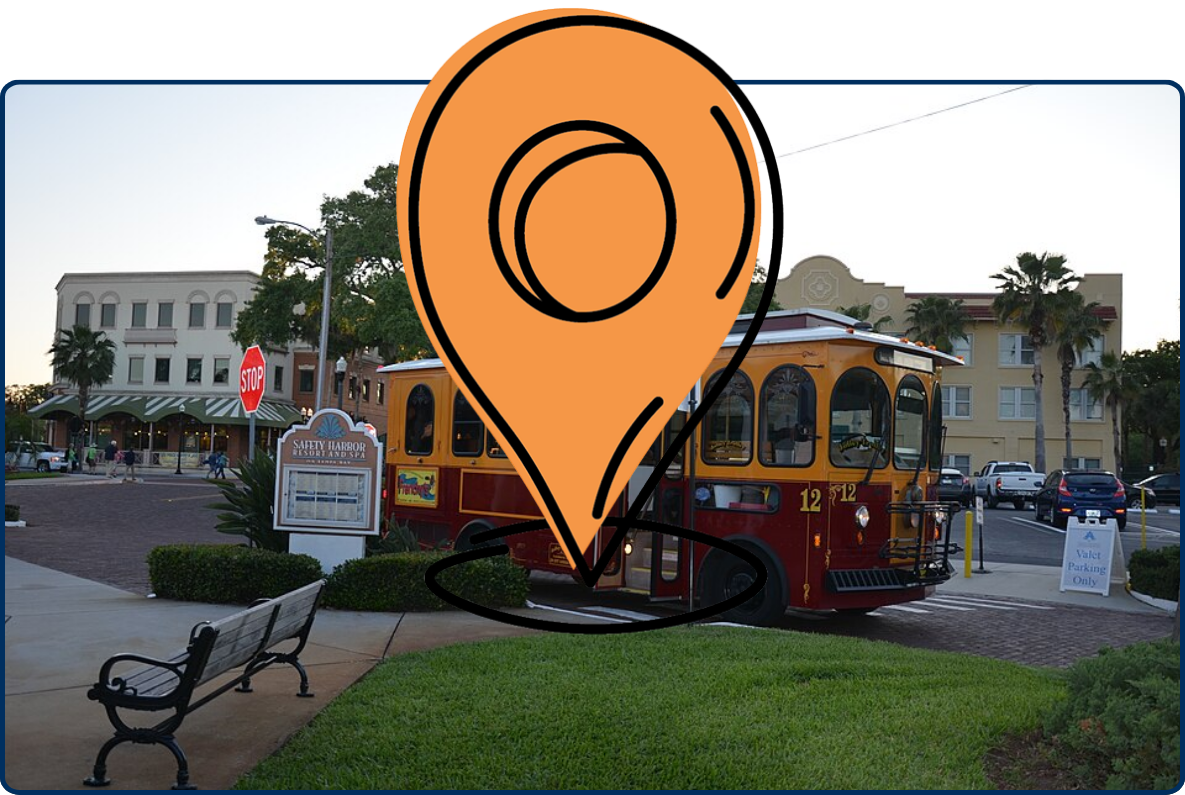 Street scene in Safety Harbor, Florida, featuring a red-and-yellow trolley bus, landscaped lawn, benches, palm trees, and historic-style buildings in the background.
