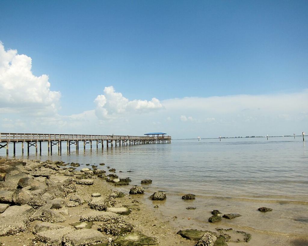 Safety Harbor waterfront in Florida, with a long wooden pier extending over calm bay waters and a rocky shoreline beneath a bright blue, partly cloudy sky.