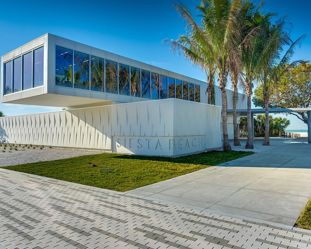Siesta Key Beach Pavilion in Sarasota, Florida, featuring modern white architecture, large glass windows, palm trees, paved walkways, and a clear blue sky.