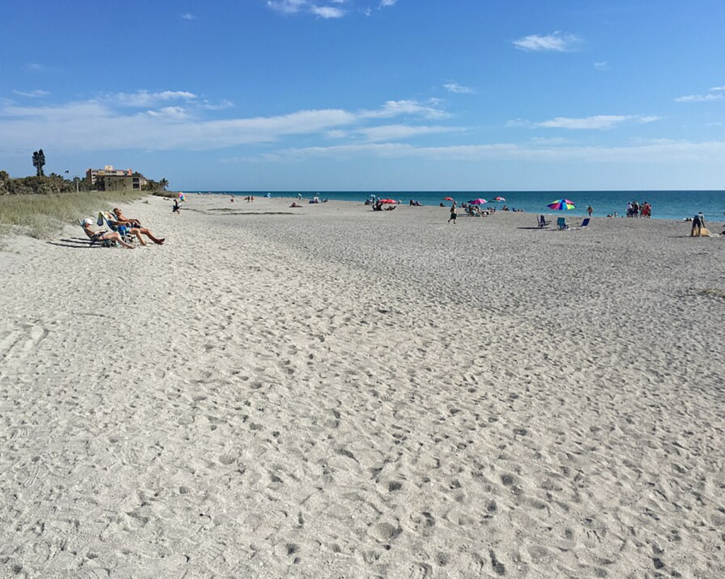 Turtle Beach on Siesta Key, Florida, with a wide sandy shoreline, scattered beachgoers, colorful umbrellas, and calm blue Gulf waters under a partly cloudy sky.
