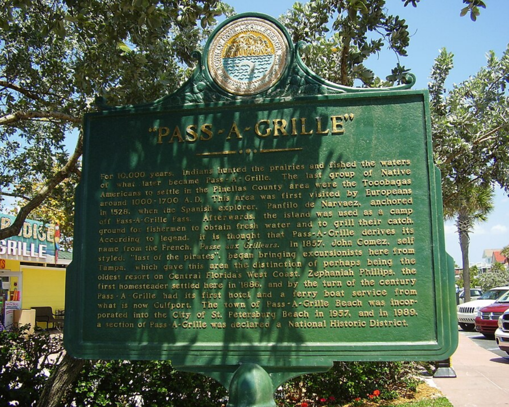 Historic Pass-a-Grille marker in St. Pete Beach, Florida, featuring a large green plaque with gold lettering surrounded by leafy trees and nearby storefronts.