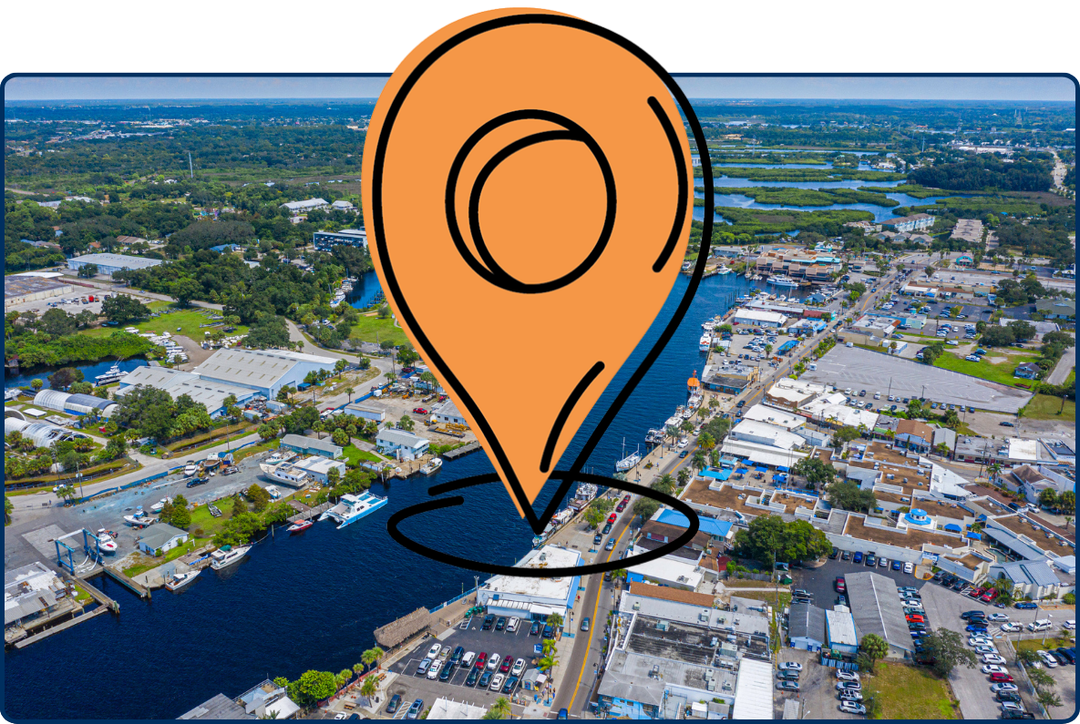 Aerial view of Tarpon Springs, Florida, showing a wide waterfront channel lined with marinas, boats, commercial buildings, roads, and surrounding wetlands beneath a partly cloudy blue sky.