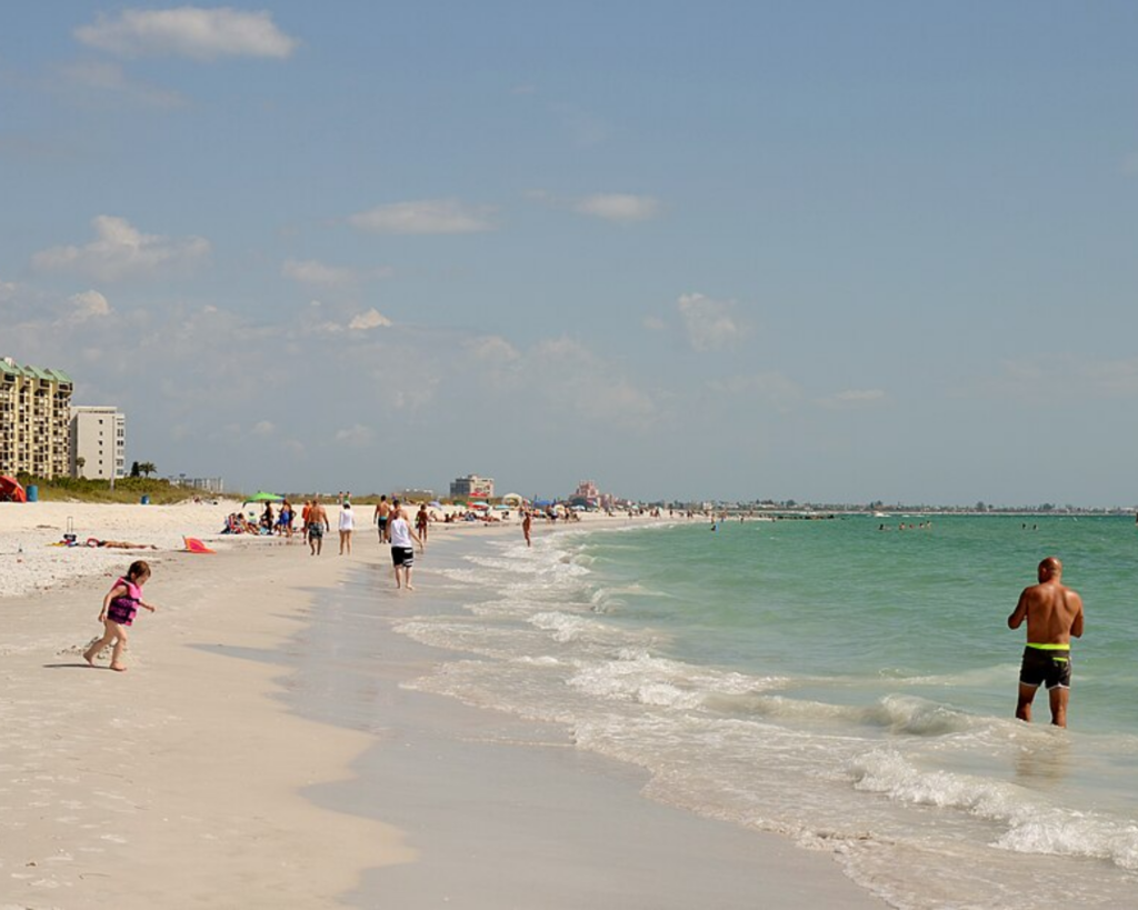 Treasure Island Beach, Florida, with a wide sandy shoreline, turquoise Gulf waters, scattered beachgoers, and beachfront buildings under a lightly clouded sky.