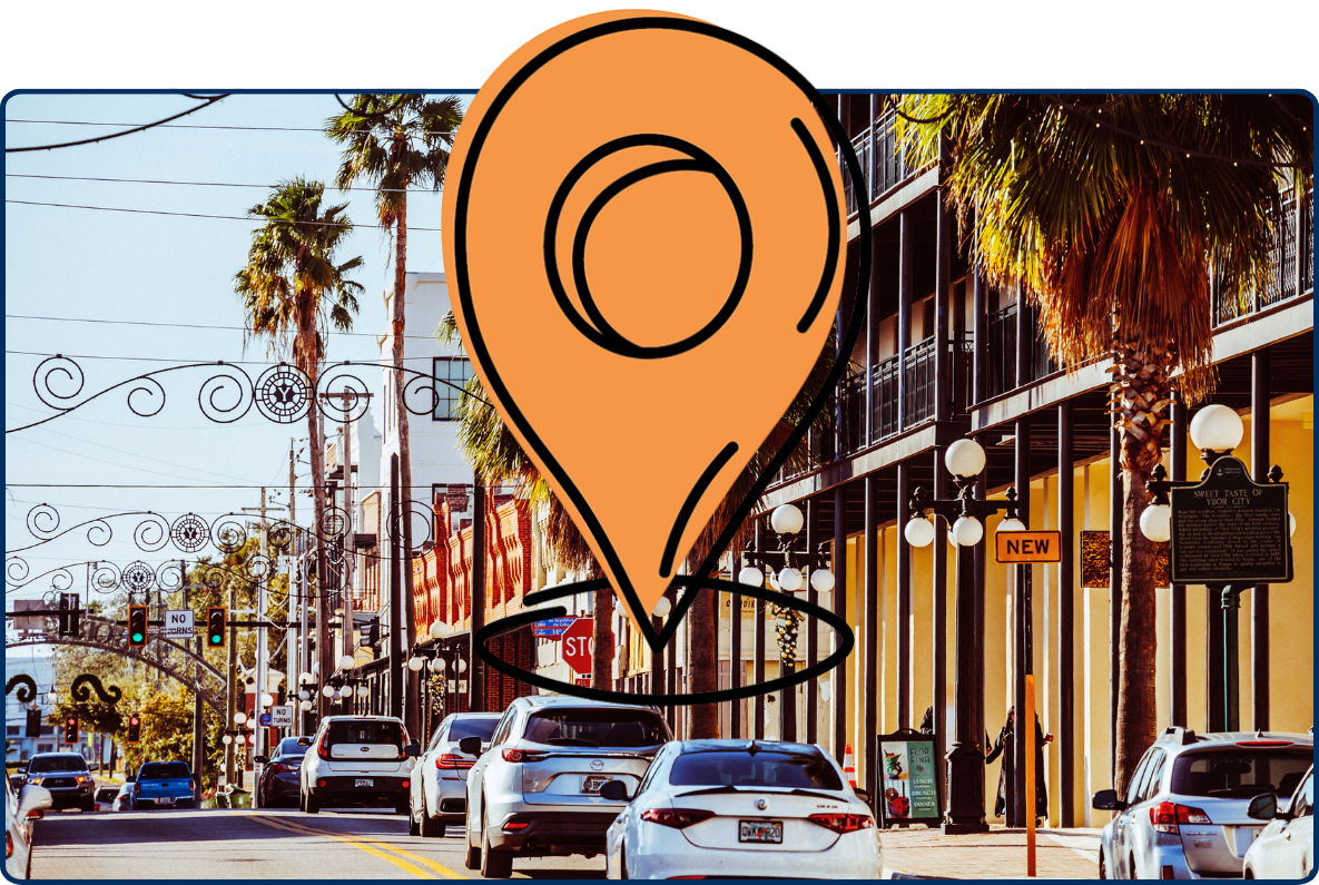 Street scene in Ybor City, Tampa, Florida, with palm trees, historic buildings, decorative street arches, parked cars, and storefronts along a sunny urban road.